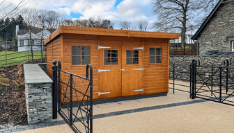 Large timber pent-roof garden shed with three doors and small windows, positioned beside a stone wall and black metal gate.